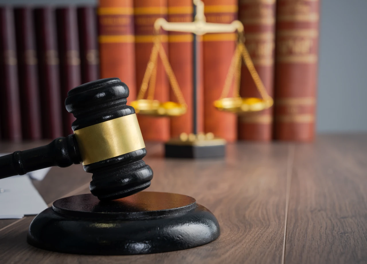 Richterhammer auf einem Holztisch, im Hintergrund eine goldene Justitia-Waage und rote Gesetzesbücher. © Daniel Tadevosyan/Shutterstock.com
