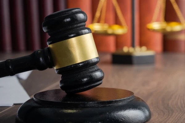 Richterhammer auf einem Holztisch, im Hintergrund eine goldene Justitia-Waage und rote Gesetzesbücher. © Daniel Tadevosyan/Shutterstock.com
